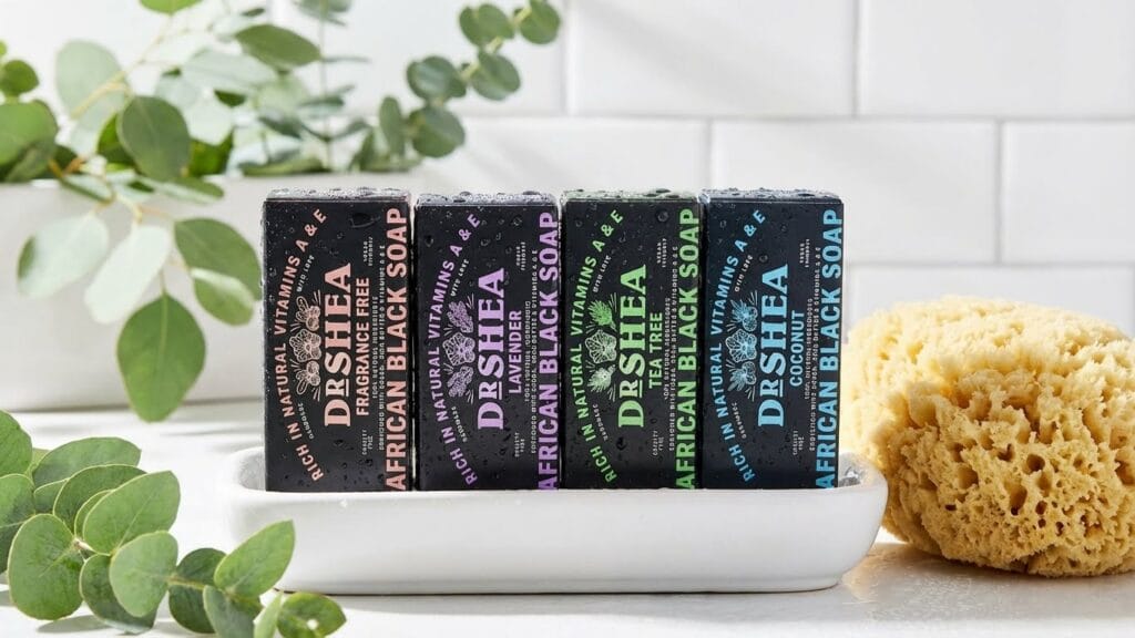 Dr Shea Store A row of four Dr Shea African Black Soap bars in Fragrance Free, Lavender, Tea Tree, and Coconut variants, displayed on a white soap dish with a natural sea sponge and green eucalyptus leaves.